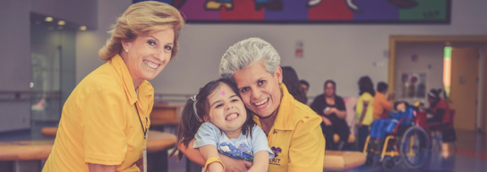 Dos voluntarias abrazando a una niña y sonriendo a la cámara