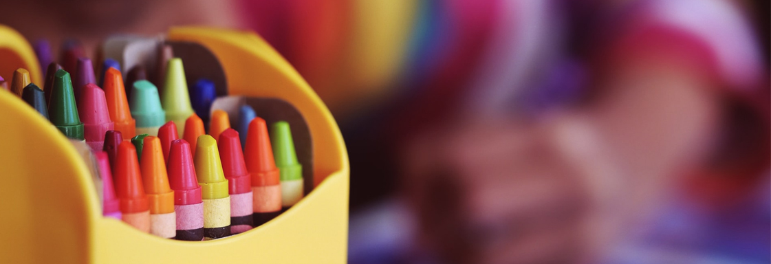 Foto de una caja de crayolas en primer plano, al fondo se ve la imagen borrosa de un niño dibujando