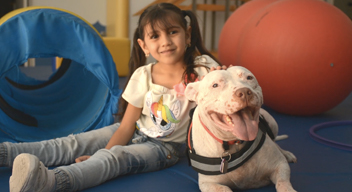 Fotografía de niña sonriendo junto a un perro blanco, sobre tapete de rehabilitación